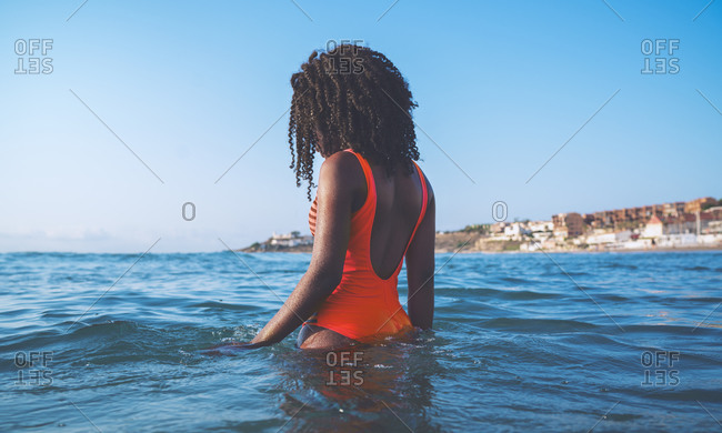 Side view of unrecognizable African American female tourist with curly hair in stylish red swimsuit standing in waving sea against cloudless blue sky