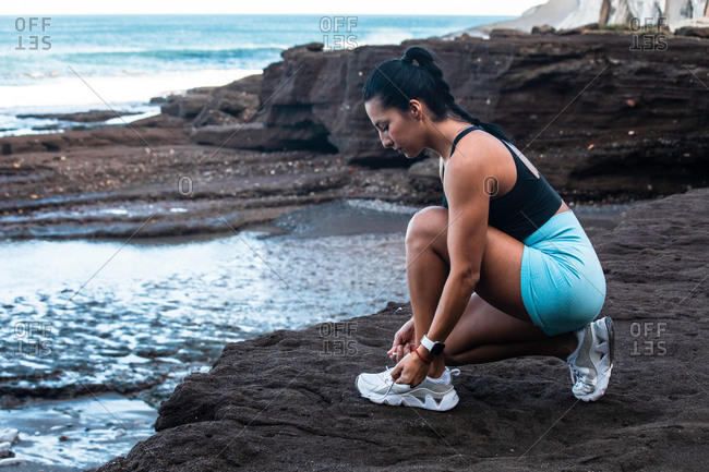 Side view of fit ethnic female athlete tying shoelaces on sneakers while sitting on seashore and preparing for workout