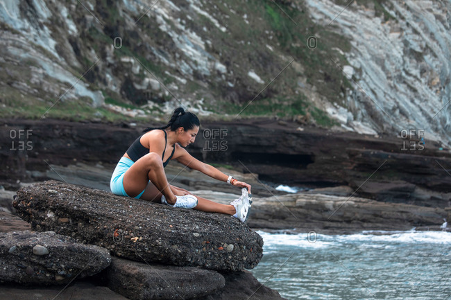 Side view of fit female athlete sitting on rock on beach and stretching legs while doing forward bend exercise during workout