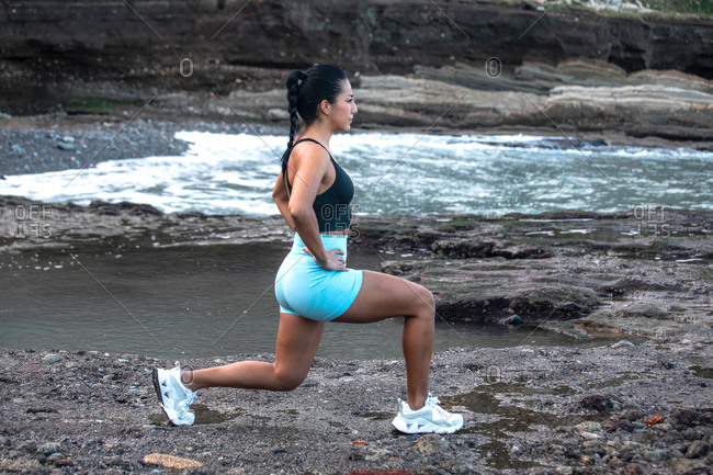 Side view of young determined athletic female doing lunges during active training on rocky seashore while looking away