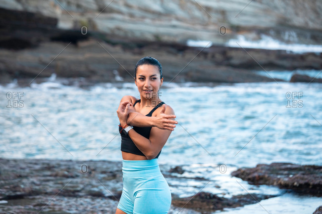 Concentrated fit female in sports bra standing in seashore and warming up while stretching arms before training and looking away