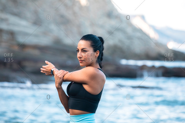 Concentrated fit female in sports bra standing in seashore and warming up while stretching arms before training and looking away