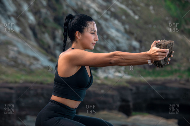 Side view of glad female athlete in sportswear doing squat exercise with rock while training in nature in summer
