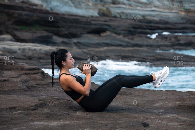 Side view of strong female athlete doing abs workout with heavy stone while training on rocky beach near sea