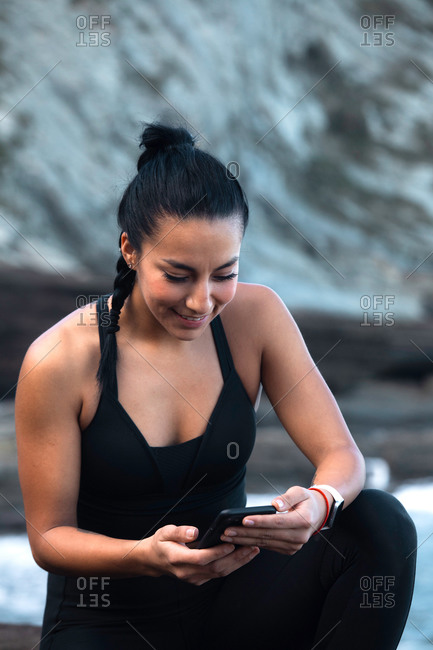 High angle side view of athletic female in activewear leaning on knee on beach near sea and surfing Internet on smartphone
