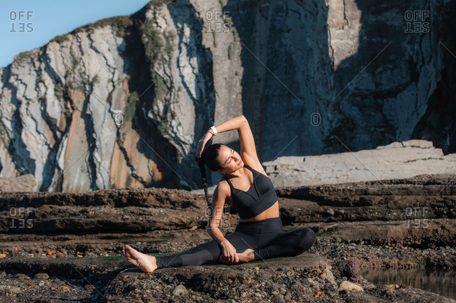 Positive female athlete warming up body and doing side bends while stretching arms before workout on seashore and looking at camera