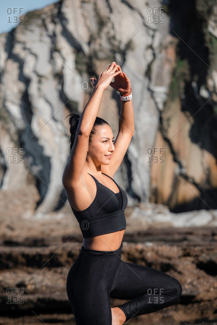 Flexible female in sportswear balancing on leg in Vrksasana with raised arms while doing yoga in mountains in summer