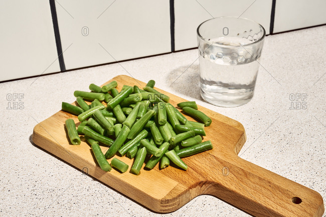 High angle pile of chopped green beans on wooden cutting board placed on table near glass of water in kitchen