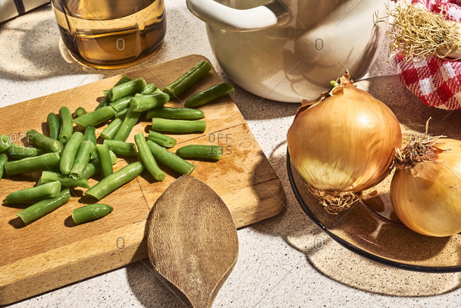 From above of fresh chopped green beans scattered on wooden cutting board placed on table near utensils and whole onions