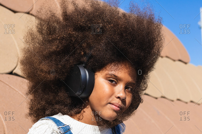 Teen black hipster girl with afro hairstyle listening to music through wireless headphones and looking at camera while resting on street