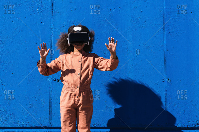 Unrecognizable African American girl in trendy suit and VR headset touching air while exploring virtual world against blue wall on street