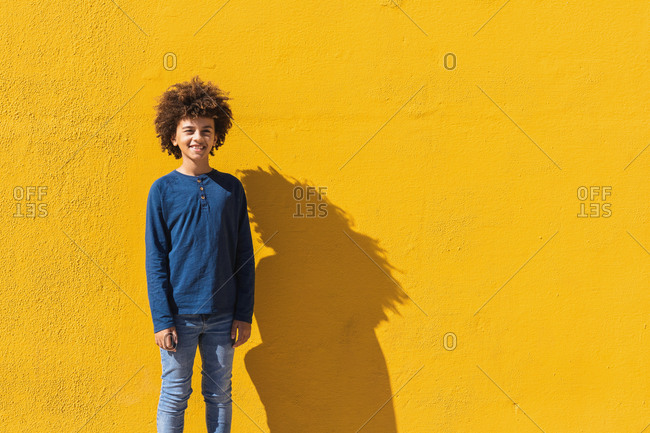 Positive male African American teenager with Afro hair looking at camera while standing against vivid yellow wall in sunlight