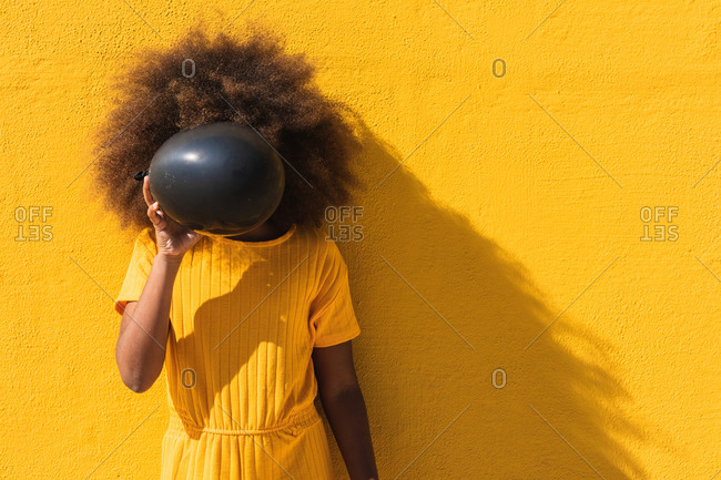 Unrecognizable curly haired teen African American girl in yellow shirt covering face with black balloons while standing against bright yellow wall
