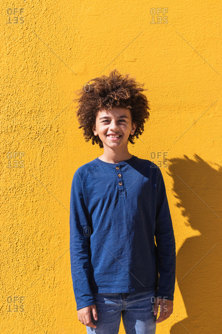 Positive male African American teenager with Afro hair looking at camera while standing against vivid yellow wall in sunlight