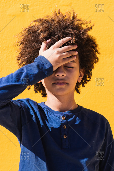 Disappointed curly haired African American teen boy with eyes closed slapping forehead while standing against bright yellow wall