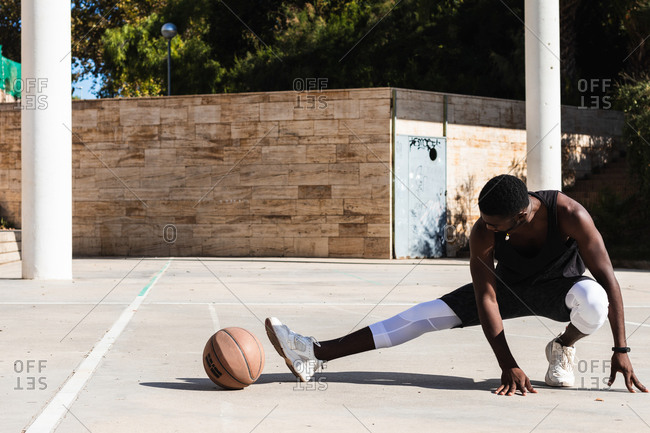 Fit African American male basketball player warming up legs and doing side lunges while preparing for training on sports ground in summer
