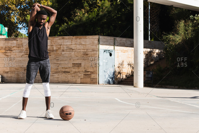 Fit African American male basketball player warming up stretching while preparing for training on sports ground in summer