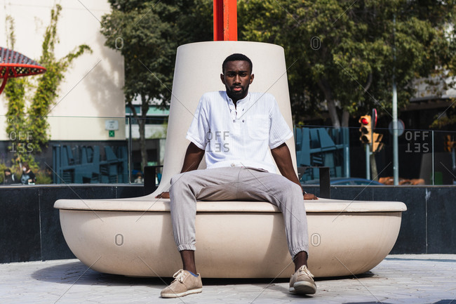 Confident African American male in summer outfit relaxing on concrete bench in city on sunny day