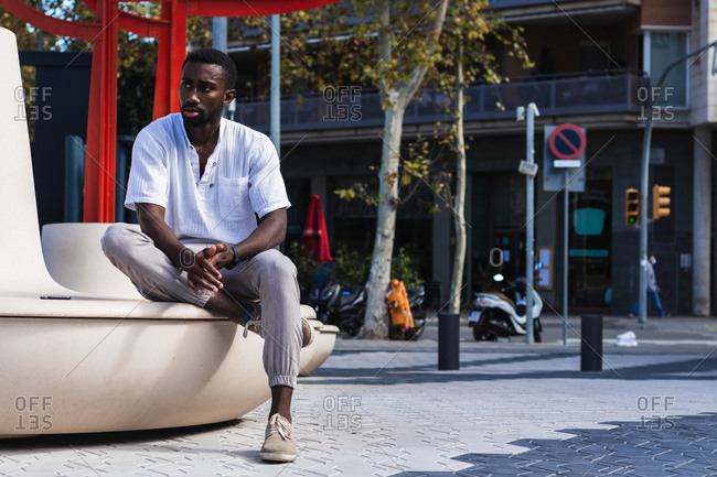 Confident African American male in summer outfit relaxing on concrete bench in city on sunny day