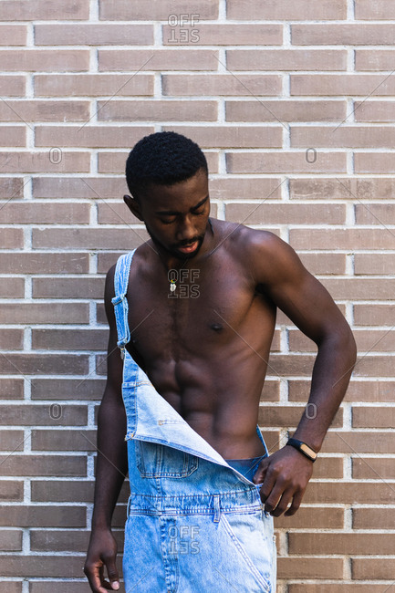 African American male with muscular naked torso standing in denim overalls in city on background of brick wall and looking down