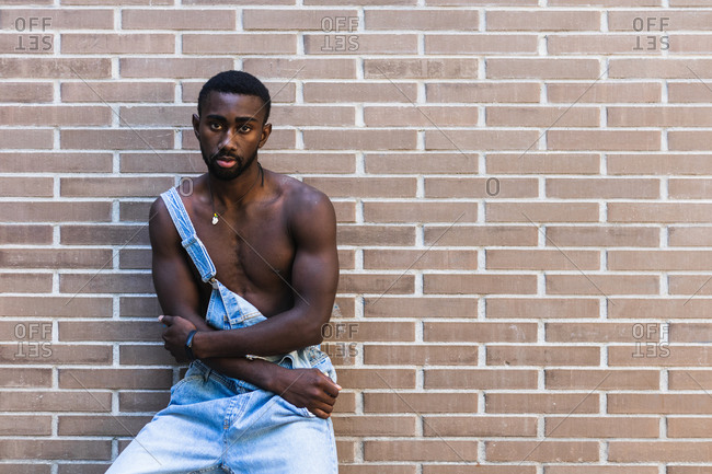 African American male with muscular naked torso standing in denim overalls in city on background of brick wall and looking at camera