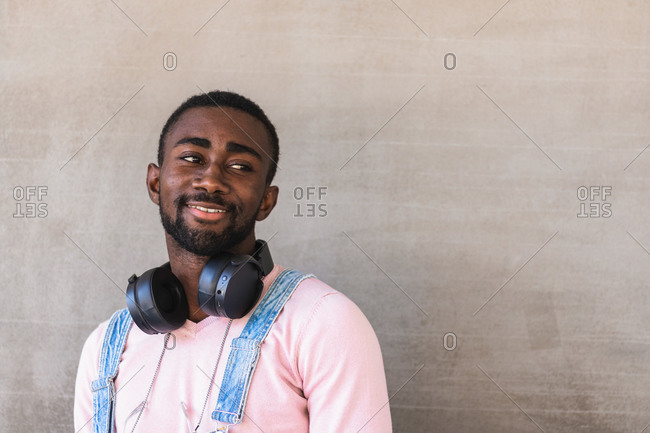 Cheerful African American male hipster in trendy clothes and with headphones on neck standing against concrete wall and looking away