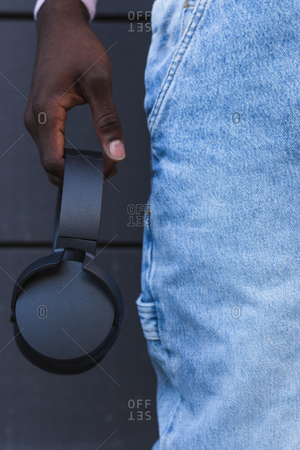 Unrecognizable crop African American male in jeans standing with black wireless headphones on street
