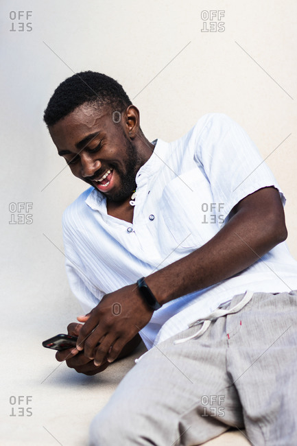 Positive cheerful African American male in casual outfit relaxing using smartphone on white background in studio and laughing