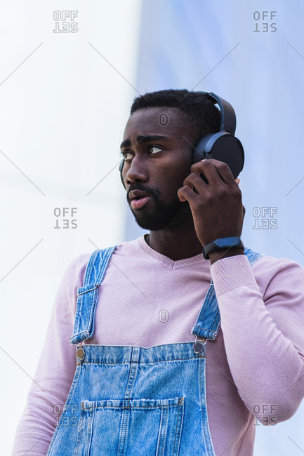 Cool African American male hipster in trendy outfit standing on street and listening to music in wireless headphones