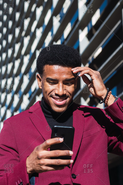 From below of happy young black guy in trendy suit smiling and touching forehead while reading good news on smartphone standing on street near contemporary building
