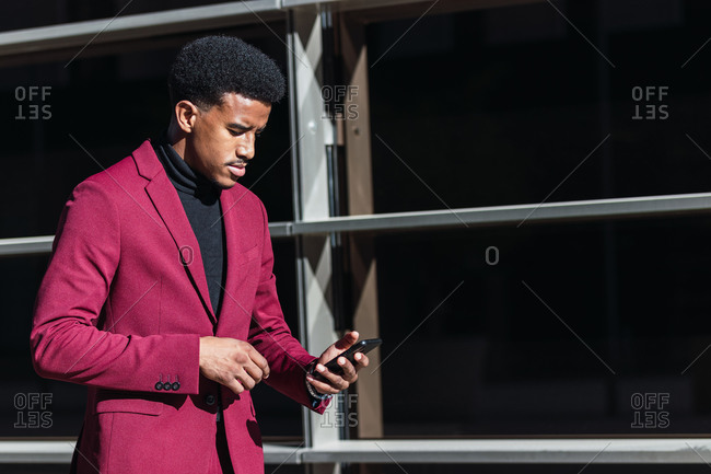 Young black guy in trendy suit smiling and touching forehead while reading good news on smartphone standing on street near contemporary building