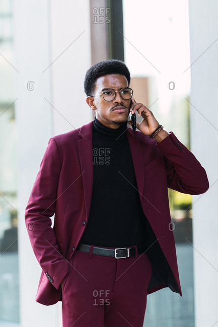 Serious young African American male manager in stylish suit and eyeglasses discussing business issues on smartphone and looking away