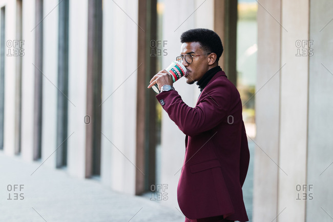 Side view of self assured young ethnic male entrepreneur in fashionable suit and eyeglasses drinking coffee from disposable cup during break on street
