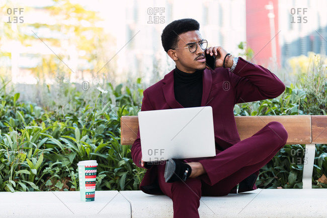 Pensive young African American male freelancer in stylish outfit and eyeglasses sitting on bench in city park and having phone conversation during remote work on laptop