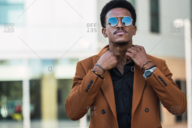 Self assured African American male wearing stylish jacket and sunglasses adjusting collar of shirt while standing in city and looking at camera