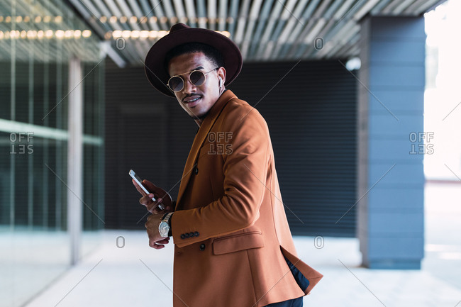 Content African American male in trendy hat and clothes walking along urban street and browsing mobile phone