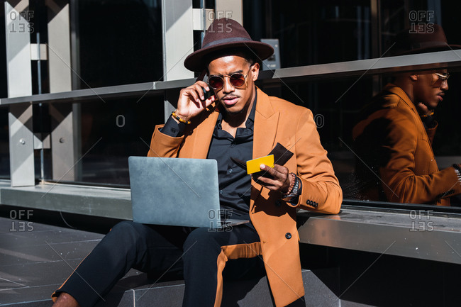 Serious African American male wearing trendy outfit and accessories sitting in street with laptop and plastic card and making purchase online