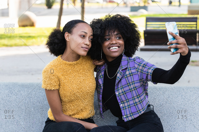 Cheerful young African American female friends hugging and taking selfie on mobile phone while spending time together on the street