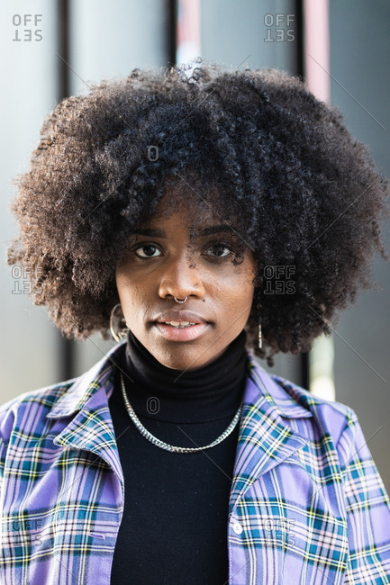 Modern young African American female with curly hair and piercing dressed in stylish checkered jacket looking at camera