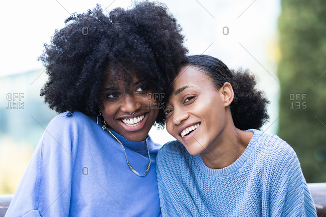 Cheerful young African American female friends with curly hair wearing similar lilac clothes cuddling together while resting on bench on urban street