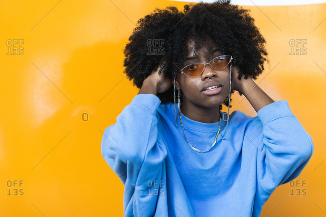 Stylish young African American female with curly hair in trendy sunglasses looking at camera while standing against yellow wall