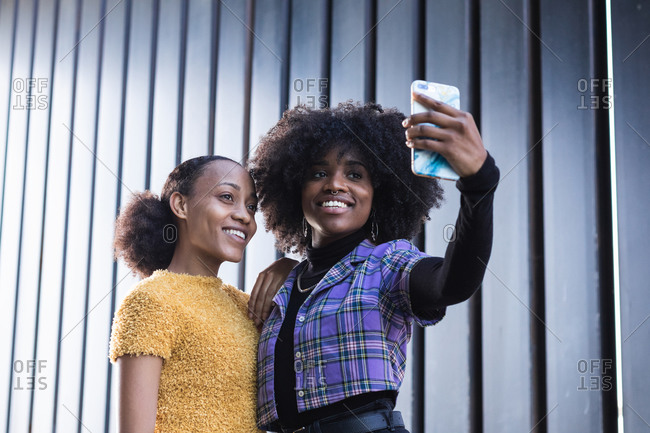 Cheerful young African American female friends hugging and taking selfie on mobile phone while spending time together on the street