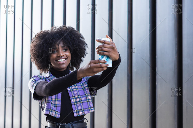 Confident smiling millennial African American female in stylish casual wear taking selfie on mobile phone while standing against wall of modern building