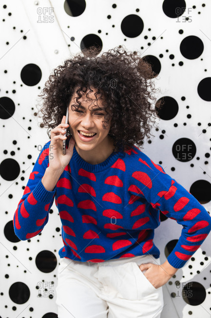 Positive African American female with curly hair and in stylish clothes standing in modern urban district and messaging on smartphone