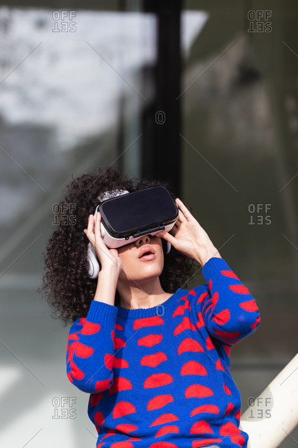 Excited female wearing modern VR glasses experiencing virtual reality while riding roundabout on playground with raised arms