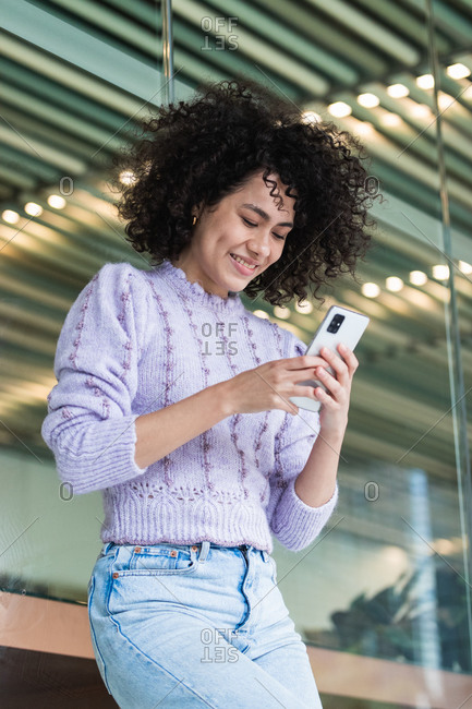 Carefree black female with Afro hairstyle standing near glass wall of modern building and having conversation on smartphone while touching hair and looking away