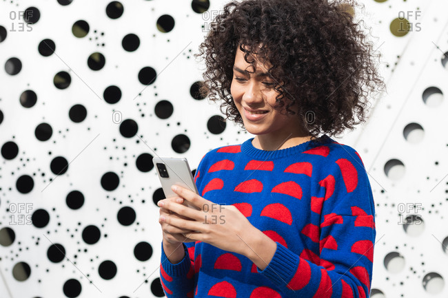 Positive African American female with curly hair and in stylish clothes standing in modern urban district and messaging on smartphone
