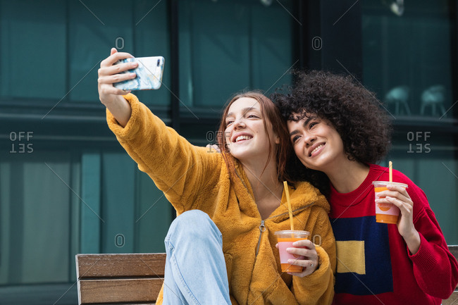 Content multiracial female best friends with takeaway juice cuddling on bench on street and taking selfie on smartphone at weekend