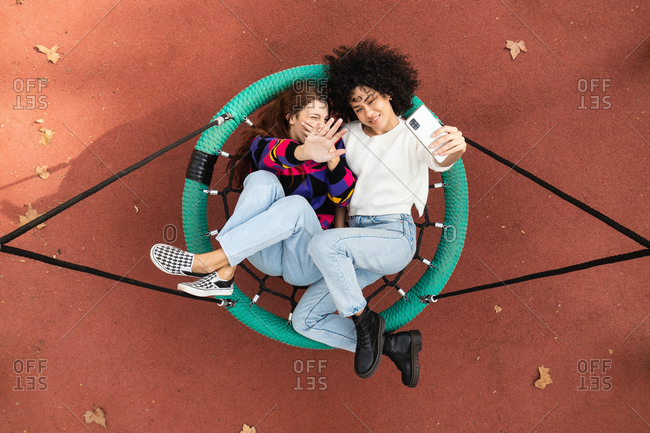 Top view of multiracial female friends lying on net swing on playground and taking selfie on smartphone while spending time together and having fun