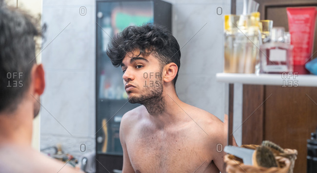 Shirtless young ethnic unshaven man looking at reflection in mirror during morning hygienic procedure in bathroom at home